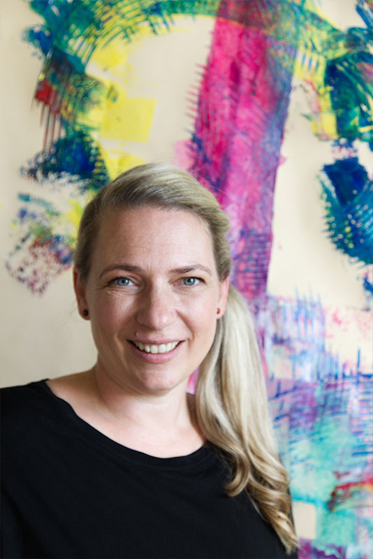 Romy Feld trägt mit einem Künstlerspachtel Farbe auf eine große Leinwand auf