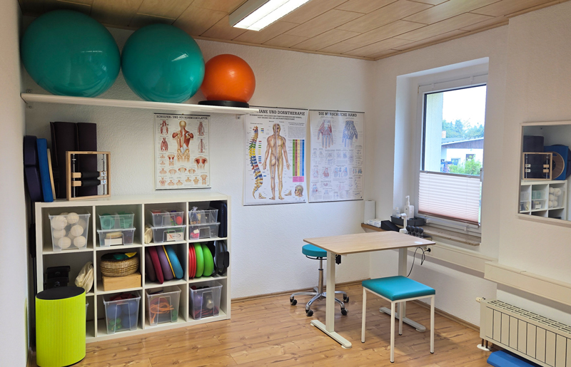 Praxisraum mit Sportgeräten, Gesundheitspostern, einem Tisch und zwei Stühlen